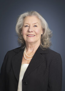Portrait of 2023-2024 District Governor Dr. Kathy Culbertson. She is in professional attire with a white blouse under a dark gray suit coat. She is wearing a double strand of pearls and is against a gray backgrounf.