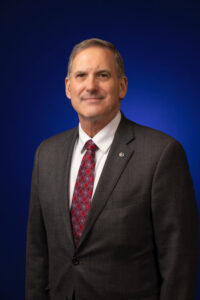 Portrait of 2024-2025 District Governor Bobby Quinten. He is in business professional attire with a white shirt, red tie, and dark gray suit coat against a blue background.