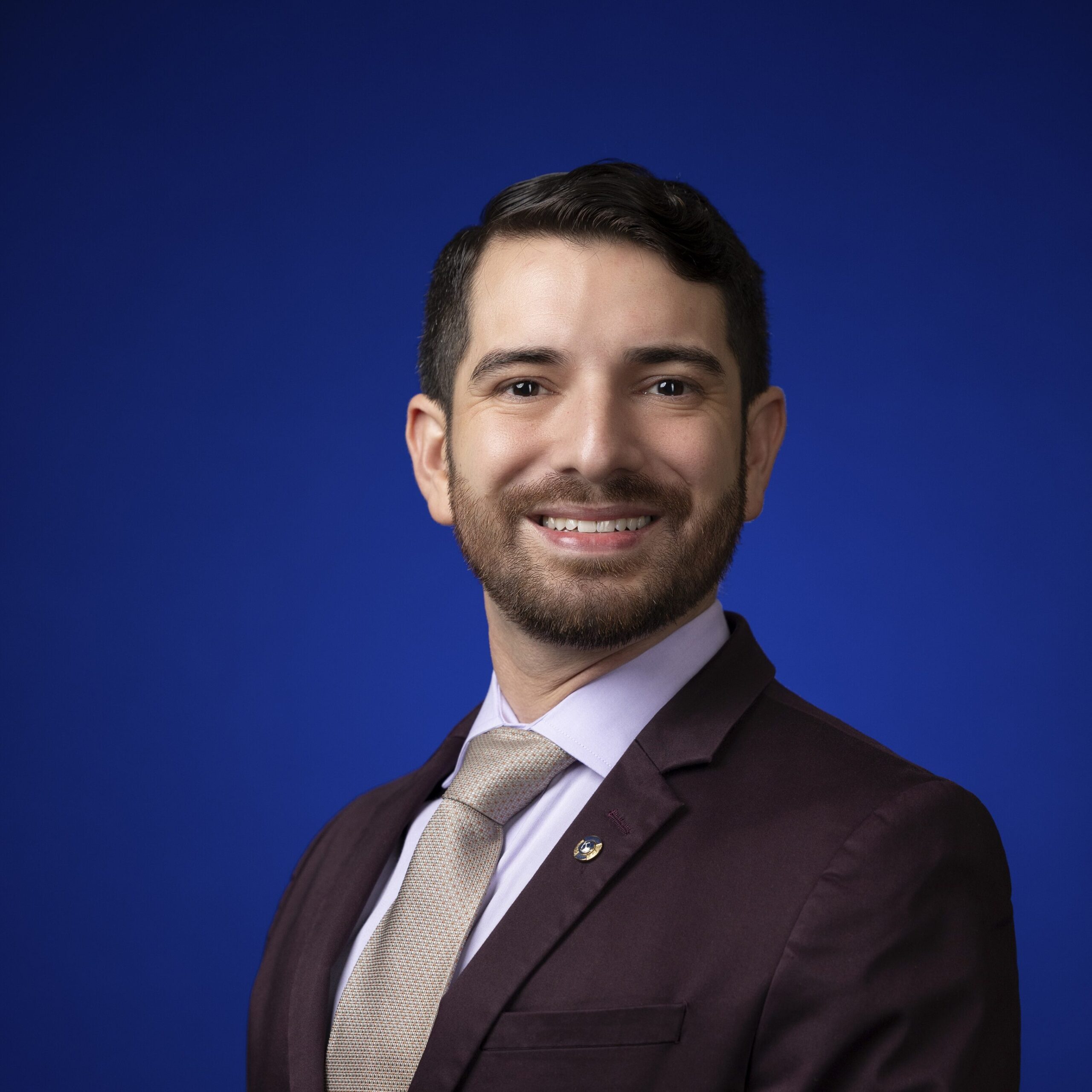 Portrait of 2025-2026 District Governor John Cano. He is in business professional dress with a white shirt, gold tie, and eggplant colored blazer against a blue background.